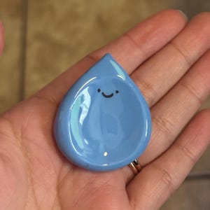 Op de afbeelding: Een lichtblauwe, druppelvormige keramische schaal met een lachend gezicht. De schaal heeft een glanzende afwerking en een kleine, ondiepe kom. Het ontwerp is eenvoudig en grillig, perfect voor kleine items.
