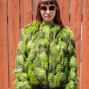 May include: A vibrant green faux fur jacket with black accents. The jacket has a shaggy texture and is open in the front. The background is a weathered red wooden fence.