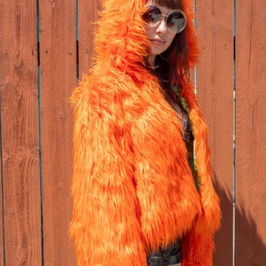 Può includere: Giacca con cappuccio in finta pelliccia arancione brillante. La giacca ha una lunga pelliccia folta e un cappuccio. La persona indossa occhiali da sole. Lo sfondo è una staccionata di legno.