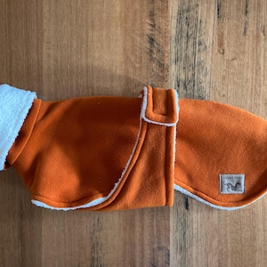 May include: Orange dog coat with white fleece lining and trim. The coat features a rectangular label with a dog silhouette. The coat is made of a soft material and has a buckle closure.