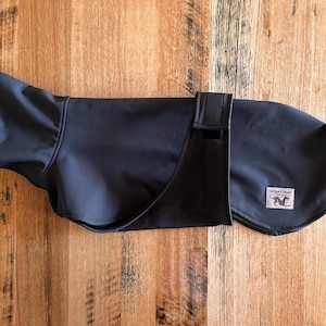 May include: A dark grey dog coat with a curved design and a rectangular patch with the text "Gadget Gear" and a dog silhouette. The coat has a strap closure and is laid flat on a wooden surface.