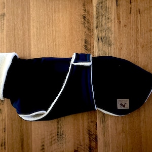 以下が含まれることがあります： 白くふわふわした内側とトリムが付いた、ネイビーブルーの犬用コート。コートにはベルクロ留めがあり、犬のシルエットが入った小さな長方形のラベルが付いています。このコートは犬を暖かく保つように設計されています。