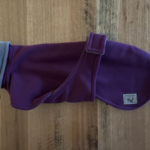May include: A purple dog coat with a grey fleece collar and lining. The coat has a flap closure with a matching purple buckle. A small label with a dog silhouette is sewn on the coat.