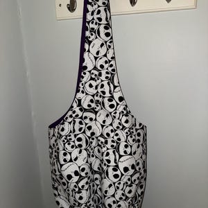 May include: A white and black tote bag featuring a repeating pattern of Jack Skellington faces. The bag has a purple interior and a long strap. The bag is hanging on a white wall with metal hooks.