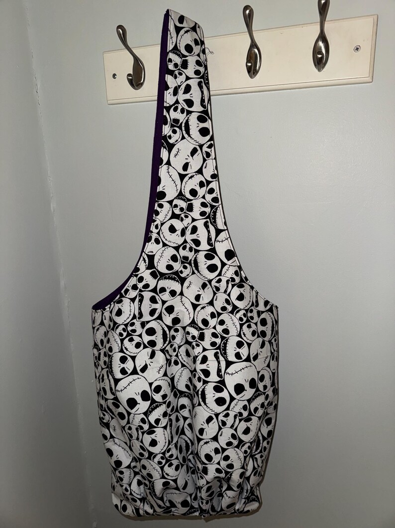 May include: A white and black tote bag featuring a repeating pattern of Jack Skellington faces. The bag has a purple interior and a long strap. The bag is hanging on a white wall with metal hooks.