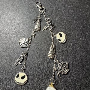 May include: A silver-toned charm keychain featuring various Halloween-themed charms. The charms include Jack Skellington faces, bats, spiderwebs, and lightning bolts. The keychain has a lobster clasp for easy attachment.