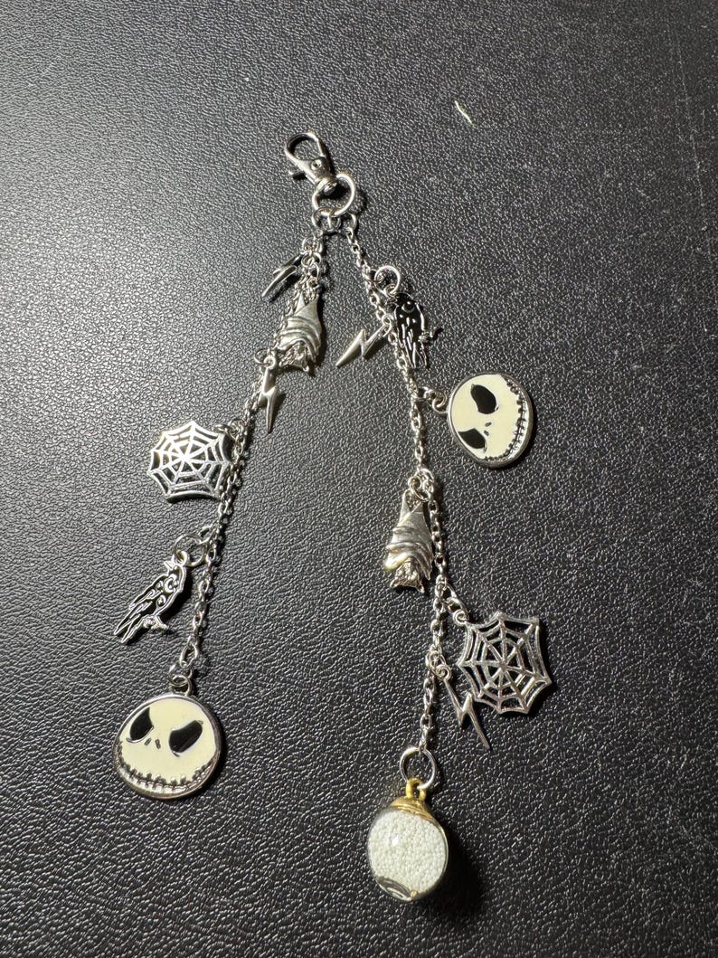 May include: A silver-toned charm keychain featuring various Halloween-themed charms. The charms include Jack Skellington faces, bats, spiderwebs, and lightning bolts. The keychain has a lobster clasp for easy attachment.
