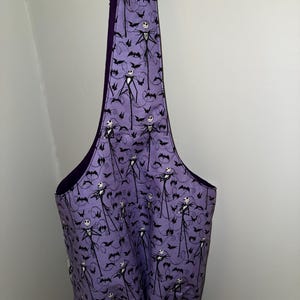 May include: A purple tote bag featuring a repeating pattern of Jack Skellington and bats. The bag has a dark purple interior and a wide, open top. The bag is hanging on a white wall.