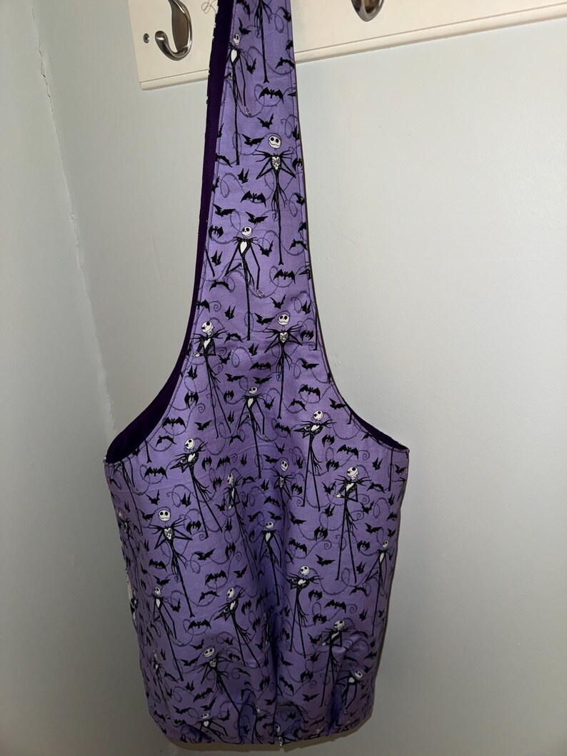 May include: A purple tote bag featuring a repeating pattern of Jack Skellington and bats. The bag has a dark purple interior and a wide, open top. The bag is hanging on a white wall.