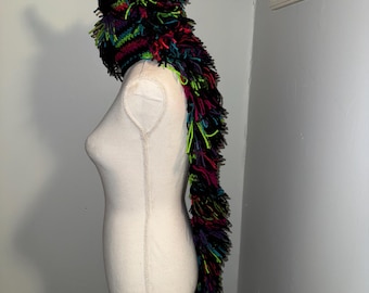 May include: A handmade, colorful monster hat with neon pink horns and a long, shaggy tail. The hat is made of black yarn with vibrant accents of green, red, and purple. The hat is displayed on a mannequin.