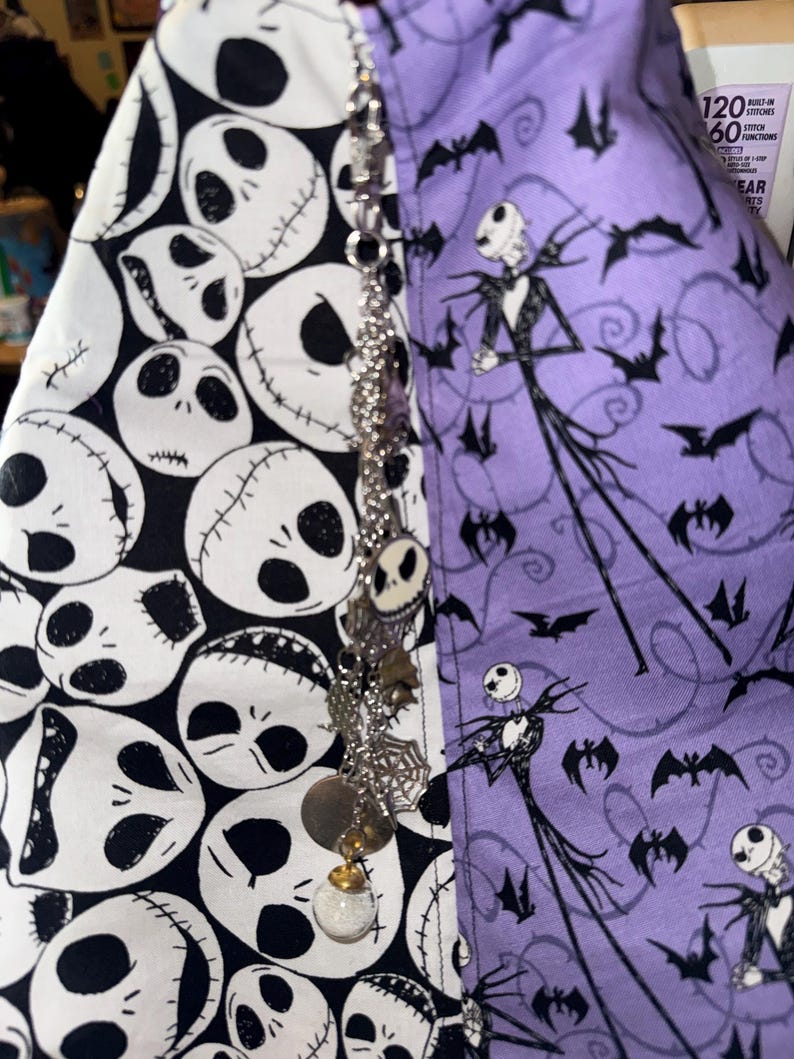 May include: A split fabric item featuring a black and white Jack Skellington pattern on one side and a purple fabric with Jack Skellington and bat silhouettes on the other. A decorative charm hangs in front.
