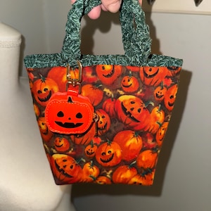 May include: A small fabric tote bag featuring a vibrant orange pumpkin pattern with jack-o'-lantern faces. The bag has dark green leafy-patterned handles and a matching orange pumpkin-shaped charm attached to a gold-toned metal clasp.