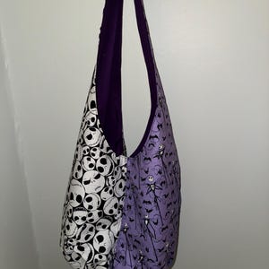 May include: A reversible shoulder bag with a Halloween theme. One side features a black and white skull pattern, while the other showcases a purple background with Jack Skellington and bat silhouettes. The bag has a purple interior and straps.