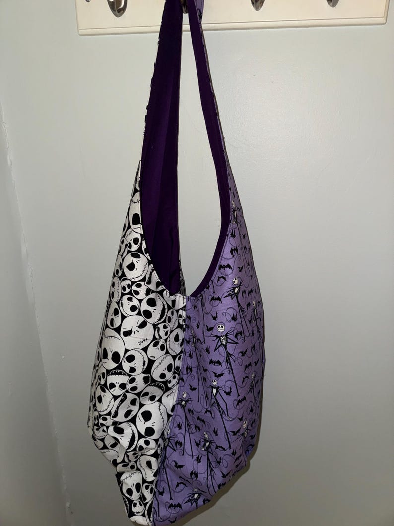 May include: A reversible shoulder bag with a Halloween theme. One side features a black and white skull pattern, while the other showcases a purple background with Jack Skellington and bat silhouettes. The bag has a purple interior and straps.