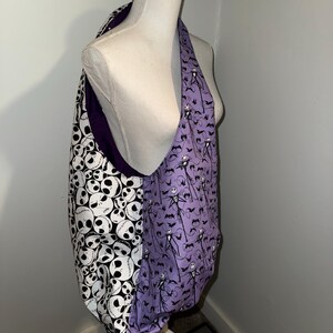 May include: A reversible tote bag with a black and white skull pattern on one side and a purple pattern featuring Jack Skellington and bats on the other. The bag is displayed on a mannequin.