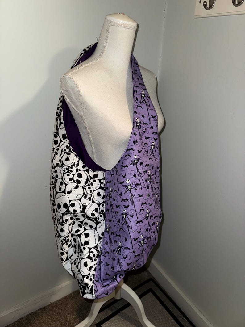 May include: A reversible tote bag with a black and white skull pattern on one side and a purple pattern featuring Jack Skellington and bats on the other. The bag is displayed on a mannequin.