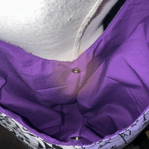May include: A tote bag with a purple interior and a black and white patterned exterior. The bag has a snap closure and a white strap. The bag is made of fabric.