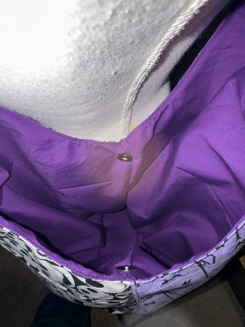May include: A tote bag with a purple interior and a black and white patterned exterior. The bag has a snap closure and a white strap. The bag is made of fabric.