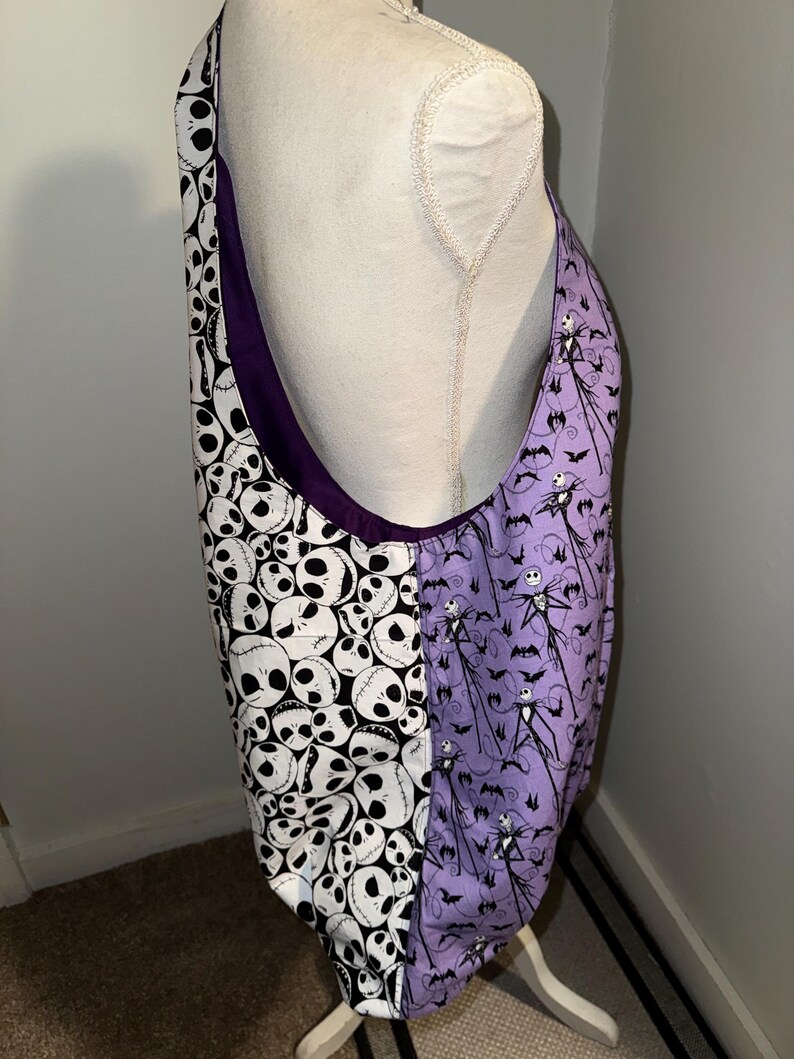 May include: A large tote bag with a purple interior and a split design. One side features a pattern of white skulls on a black background, while the other side has a purple background with Jack Skellington and bat silhouettes.