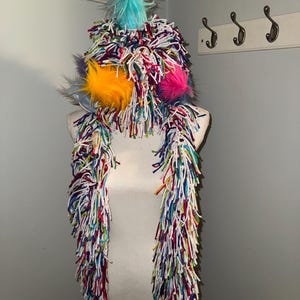 May include: A whimsical, handmade hat and scarf set. The hat features a fluffy, multi-colored design with a turquoise plume and vibrant orange and pink pom-poms. The matching scarf is long and fringed, with a rainbow of colors.