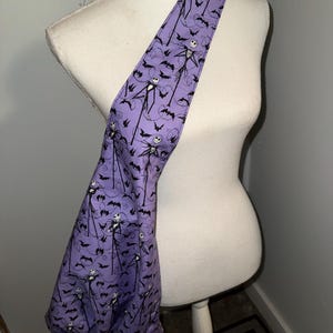 May include: A purple shoulder bag featuring a repeating pattern of Jack Skellington and bats. The bag is made of fabric and has a long strap. The bag is displayed on a white mannequin.
