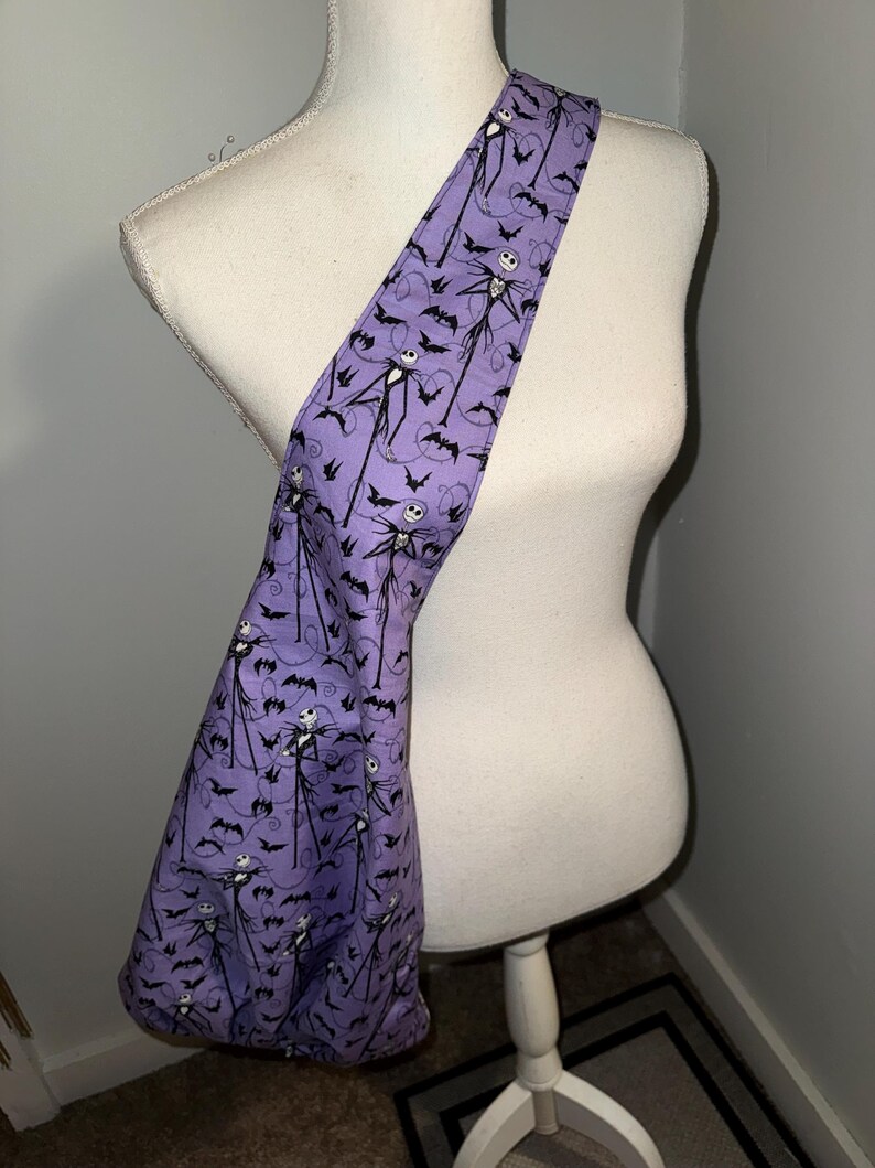 May include: A purple shoulder bag featuring a repeating pattern of Jack Skellington and bats. The bag is made of fabric and has a long strap. The bag is displayed on a white mannequin.