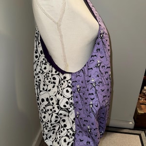 May include: A large tote bag with a purple interior. One side features a pattern of white skulls on a black background, while the other side displays a purple pattern with Jack Skellington and bats. The bag is made of fabric.