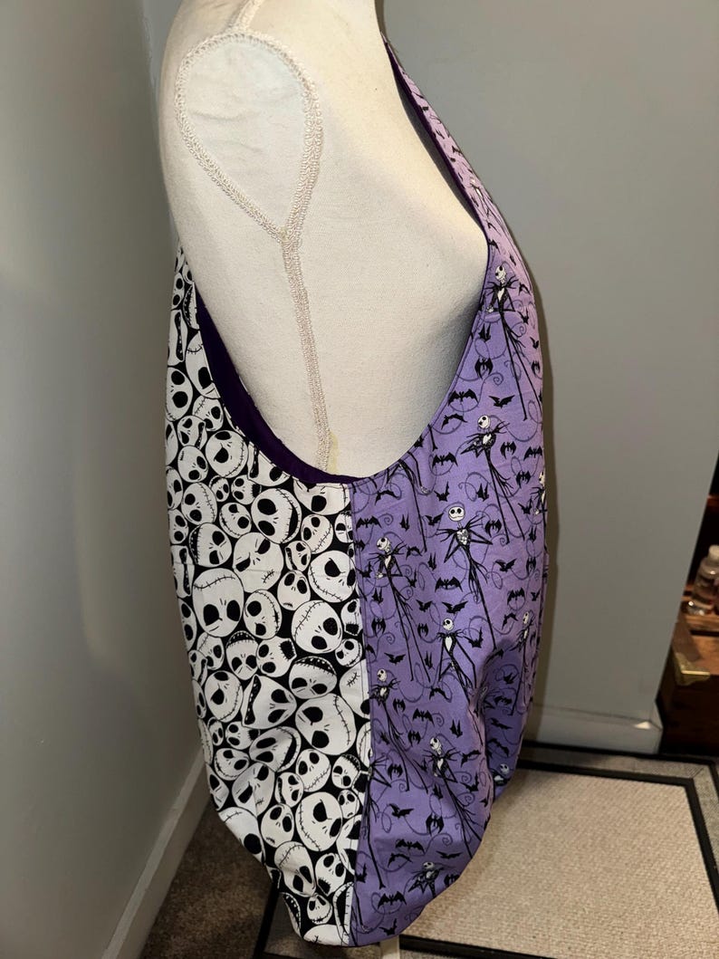 May include: A large tote bag with a purple interior. One side features a pattern of white skulls on a black background, while the other side displays a purple pattern with Jack Skellington and bats. The bag is made of fabric.