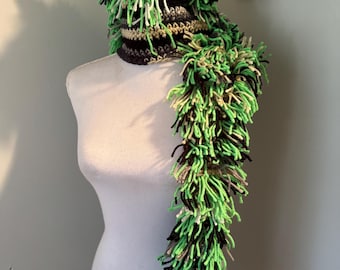 May include: A handmade, whimsical crocheted hat and scarf set. The hat features a horn and ear details, with a shaggy, green, black, and white fringe. The scarf extends down the mannequin, also with fringe. The set is displayed on a white mannequin.