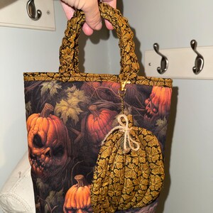 May include: A fabric tote bag featuring a dark print of orange jack-o'-lanterns and autumn leaves. The bag has a braided fabric handle and a matching pumpkin-shaped charm attached to a gold-tone metal lobster clasp hanging from the handle.