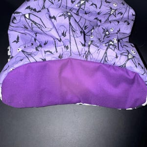 May include: A purple fabric hood with a black and white Jack Skellington pattern on the outside and a solid purple lining. The hood features a repeating pattern of Jack Skellington figures and bats.