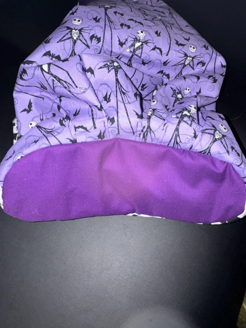 May include: A purple fabric hood with a black and white Jack Skellington pattern on the outside and a solid purple lining. The hood features a repeating pattern of Jack Skellington figures and bats.
