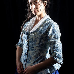 May include: A woman wearing a blue and white toile print blouse with a ruffled collar and sleeves. The blouse is buttoned up and has a white lace insert. The background is black.
