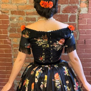 May include: A black off-the-shoulder dress with a floral pattern in red, yellow, and blue. The dress features a corset back, fringe detailing, and a velvet waistband. Orange flower hair accessories complete the look.