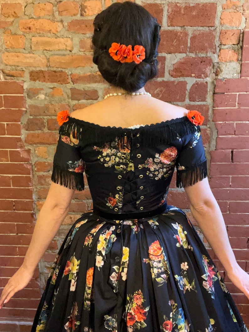 May include: A black off-the-shoulder dress with a floral pattern in red, yellow, and blue. The dress features a corset back, fringe detailing, and a velvet waistband. Orange flower hair accessories complete the look.