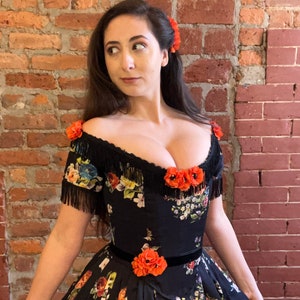 May include: A black off-the-shoulder dress with a floral pattern in various colors. The dress features a black fringe trim, orange floral accents, and a black velvet belt. A matching orange flower is in the hair.
