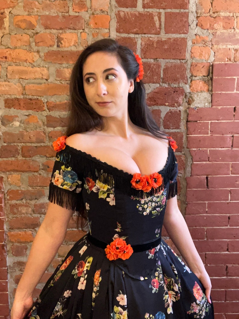 May include: A black off-the-shoulder dress with a floral pattern in various colors. The dress features a black fringe trim, orange floral accents, and a black velvet belt. A matching orange flower is in the hair.
