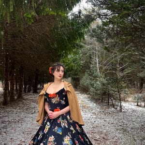 May include: A woman in a black floral print gown and a tan cape stands in a snowy forest. The dress features a sweetheart neckline and a full skirt with colorful flowers. She wears a flower in her hair.