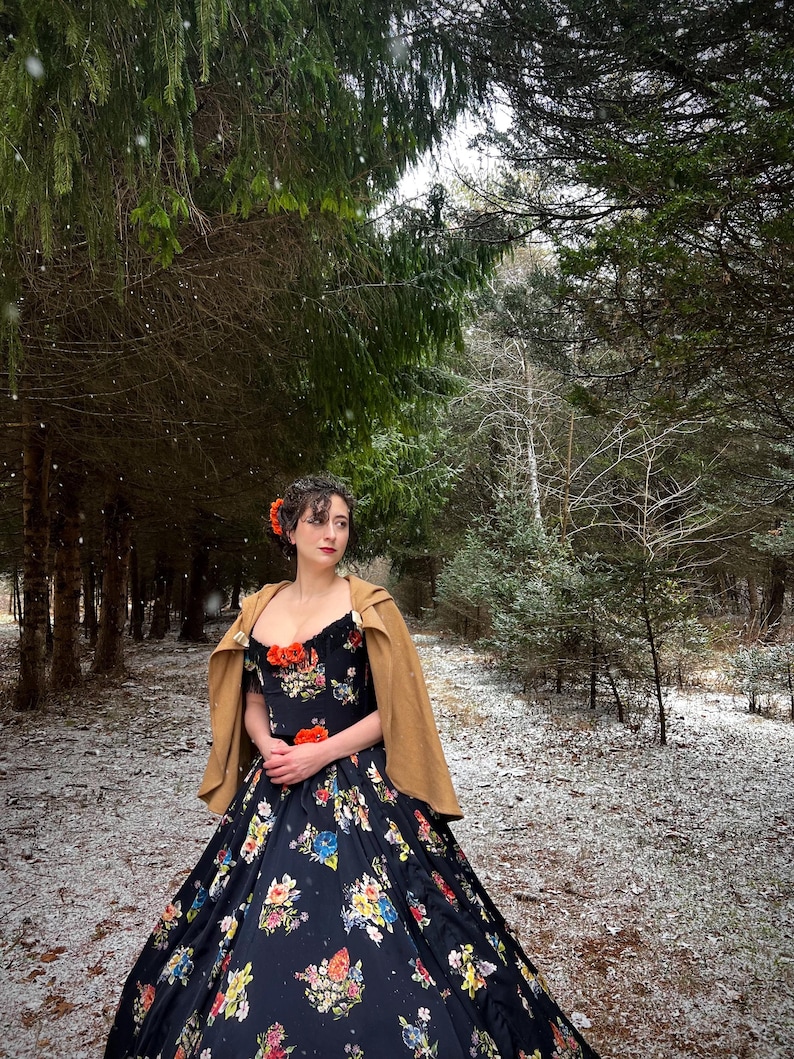 May include: A woman in a black floral print gown and a tan cape stands in a snowy forest. The dress features a sweetheart neckline and a full skirt with colorful flowers. She wears a flower in her hair.