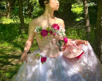 May include: A strapless, pale pink and white gown with floral detailing. The bodice is decorated with pink and red flowers, and the skirt has gold accents. The dress is worn outdoors in a forest setting.