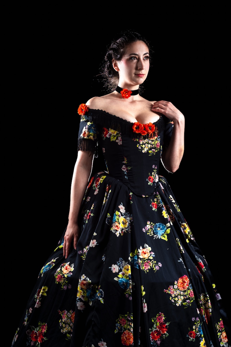 May include: A black off-the-shoulder gown with a floral pattern in red, yellow, blue, and white. The dress features a fitted bodice, a full skirt, and is adorned with red flower accents. A black choker with a red flower is also visible.