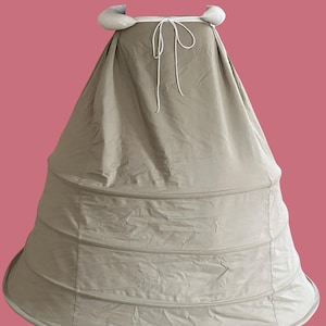 Könnte beinhalten: Ein beiger Crinoline-Unterrock mit einer breiten, glockenförmigen Silhouette. Der Unterrock hat mehrere horizontale Reifen für Struktur und Halt. Er hat einen Kordelzugverschluss und gepolsterte Akzente an der Oberseite. Der Hintergrund ist einfarbig rosa.