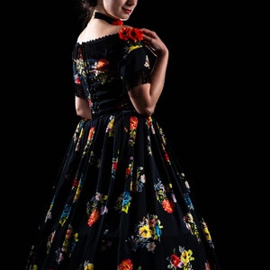 May include: A black floral print dress with a full skirt and short sleeves. The dress features a colorful floral pattern and fringe detailing on the sleeves. A red flower is pinned on the shoulder. The dress is set against a black background.