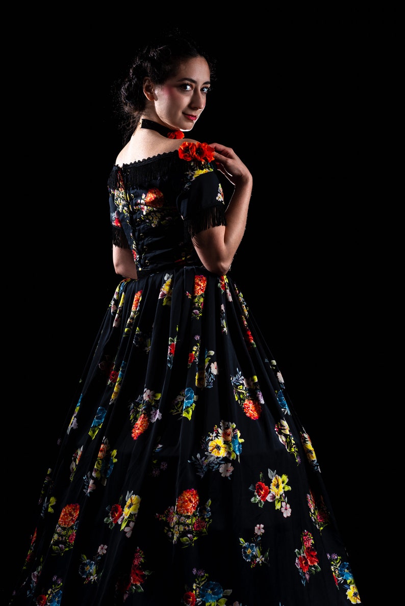 May include: A black floral print dress with a full skirt and short sleeves. The dress features a colorful floral pattern and fringe detailing on the sleeves. A red flower is pinned on the shoulder. The dress is set against a black background.