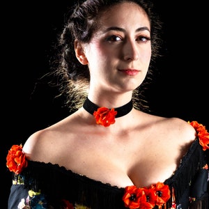 May include: A woman wearing a black velvet choker with a red flower accent. She is also wearing a black off-the-shoulder top with floral print and fringe detailing, adorned with red flower accents.