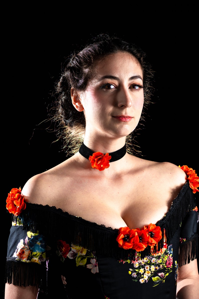 May include: A woman wearing a black velvet choker with a red flower accent. She is also wearing a black off-the-shoulder top with floral print and fringe detailing, adorned with red flower accents.