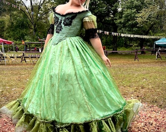 May include: A light green historical dress with a low neckline, black lace accents, and a full skirt. The dress features gold embroidery and a layered, ruffled hem. The sleeves are short and adorned with lace. The dress is suitable for cosplay or historical reenactments.