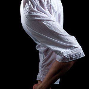 May include: White cotton bloomers featuring a high-waisted design with two small buttons at the center front waistband. These loose-fitting, knee-length trousers have gathered fabric at the waist and horizontal stitched detailing at the hem.