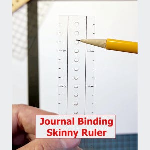 May include: A clear, skinny ruler with evenly spaced holes and numbered markings. A yellow pencil is positioned near the holes. The text "Journal Binding Skinny Ruler" is displayed in a red box at the bottom. Ideal for journal making.