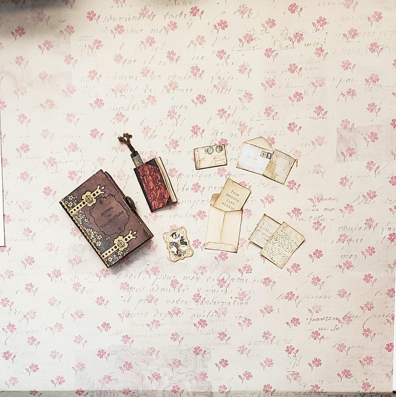 Pu&ograve; includere: Un diario in stile vintage con una copertina in pelle marrone e dettagli dorati, insieme a diverse buste in miniatura e un piccolo libro con un design floreale. Gli articoli sono disposti su uno sfondo con un delicato motivo floreale rosa.